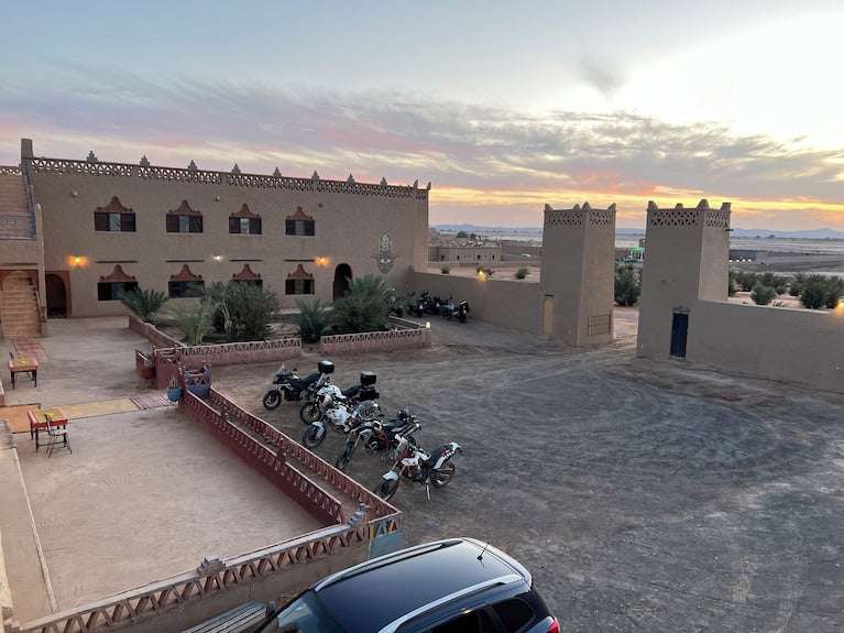 Hotel Camping Les Roses featuring traditional beige adobe buildings with decorative crenellations and a motorcycle parking area under a colorful sunset sky