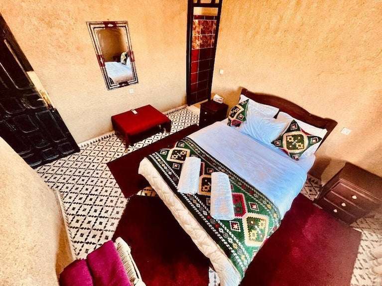 Hotel Camping Les Roses bedroom with orange textured walls, patterned tile floor, wooden bed with geometric textile throw and pillows, burgundy rug, and two towels on the bed