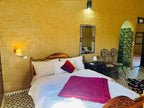 Hotel Camping Les Roses bedroom with white bedding, purple blanket and pillow, rustic wooden headboard, patterned tile floor, and earth-toned walls