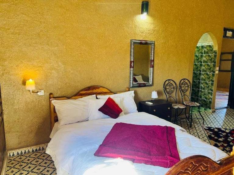 Hotel Camping Les Roses bedroom with white bedding, purple blanket and pillow, rustic wooden headboard, patterned tile floor, and earth-toned walls