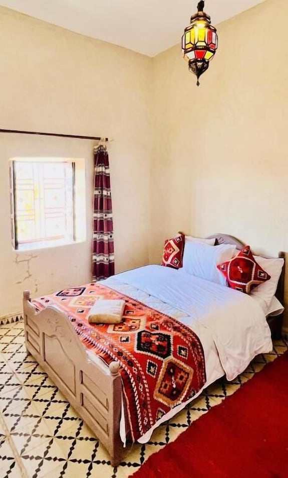 Hotel Camping Les Roses bedroom with wooden bed, white bedding, red and orange patterned blanket and pillows, tiled floor and decorative lantern