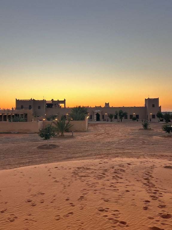 Hotel Camping Les Roses desert lodge with adobe-style buildings and palm trees at sunset with sandy foreground