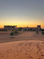 Hotel Camping Les Roses desert lodge with adobe-style buildings and palm trees at sunset with sandy foreground