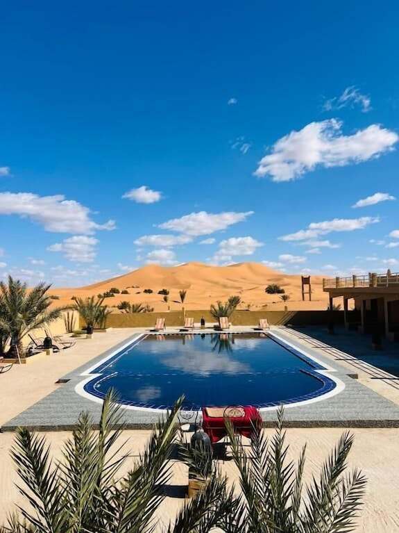 Outdoor rectangular blue swimming pool with white tiles, surrounded by palm trees and desert dunes at Hotel Camping Les Roses