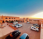 Hotel Camping Les Roses showing traditional desert-style buildings with beige walls and red accents, parked cars in a spacious courtyard under clear sky at sunset