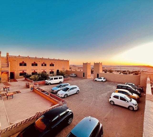 Hotel Camping Les Roses showing traditional desert-style buildings with beige walls and red accents, parked cars in a spacious courtyard under clear sky at sunset