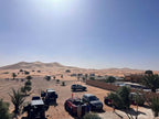 Hotel Camping Les Roses showing off-road vehicles parked on sandy desert terrain near palm trees and a compound with desert dunes in the background under clear sky