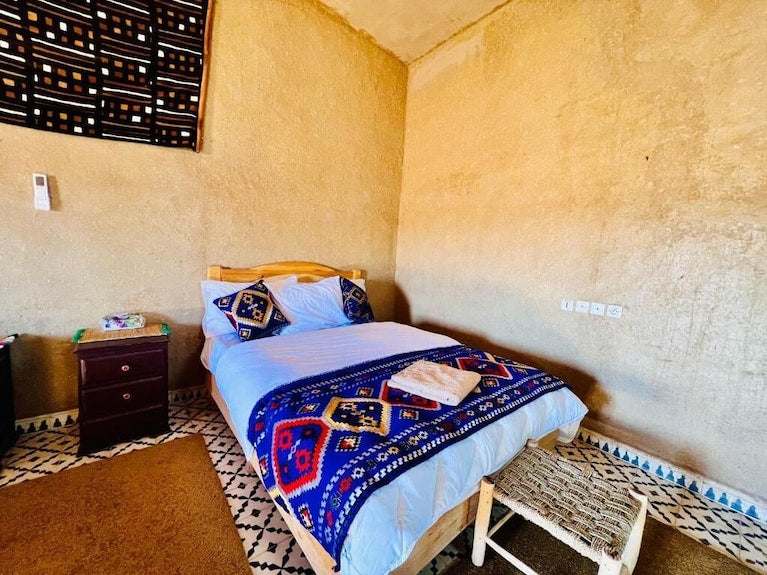 Hotel Camping Les Roses room with beige textured walls, single bed with white linens and colorful blue red patterned blanket and pillows