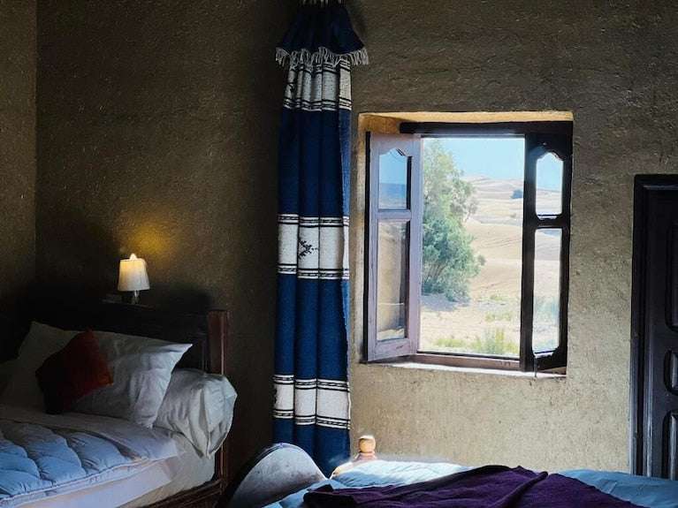 Hotel Camping Les Roses rustic bedroom with white bedding, dark wooden furniture, blue and white patterned curtain, and open window overlooking desert and greenery