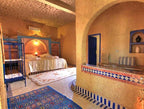 Hotel Kasbah Ksar Bicha bedroom with warm ochre walls, Moroccan tiled accents, blue metal bed frame, and patterned rugs