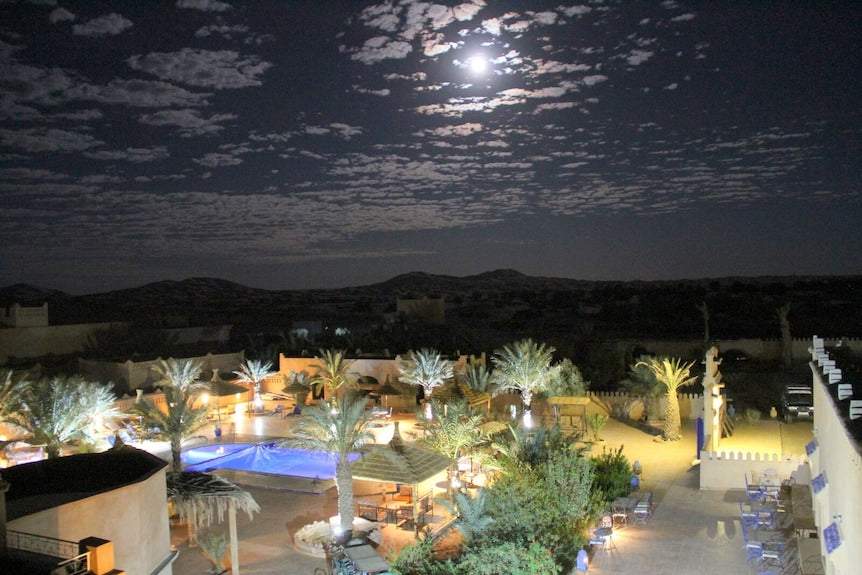 Hotel Kasbah Ksar Bicha at night with illuminated pool, palm trees, and traditional Moroccan kasbah architecture under a cloudy moonlit sky