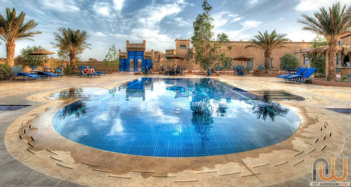 Hotel Kasbah Ksar Bicha outdoor swimming pool with blue lounge chairs, palm trees, and traditional Moroccan kasbah architecture