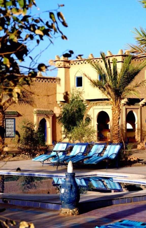Hotel Kasbah Ksar Bicha features traditional mud-brick architecture with archways, palm trees, and blue lounge chairs by a reflective pool.