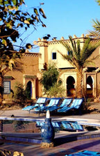 Hotel Kasbah Ksar Bicha features traditional mud-brick architecture with archways, palm trees, and blue lounge chairs by a reflective pool.