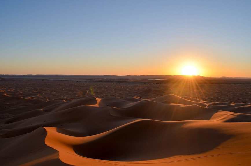 Sunset over golden sand dunes in the Moroccan Sahara Desert with soft shadows and a clear sky, showcasing Hotel Kasbah Ksar Bicha surroundings