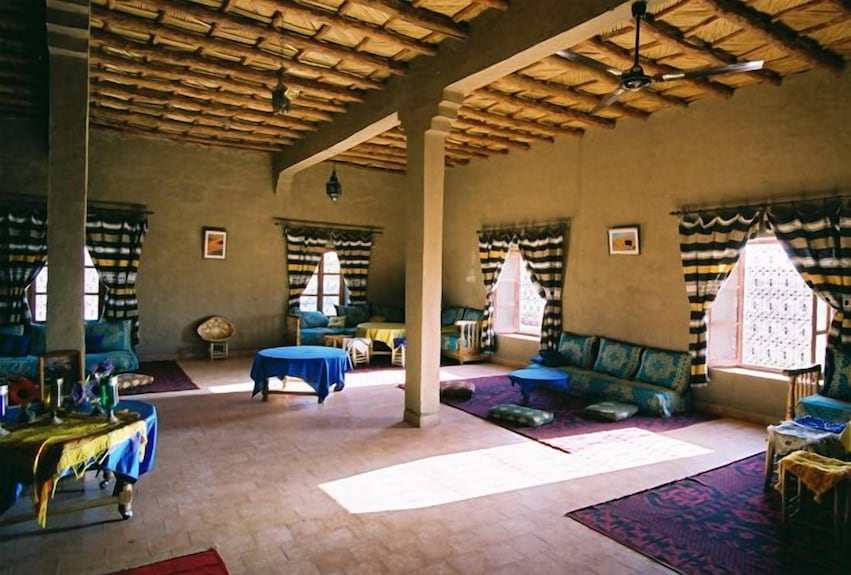 Hotel Kasbah Ksar Bicha living area with traditional Moroccan décor, blue and purple cushions, wooden windows, and bamboo ceiling