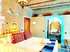 Hotel Ksar Merzouga bedroom with beige textured walls, colorful geometric-patterned pillows, white bedding, and blue tiled archway