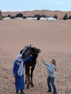 Hotel Ksar Merzouga desert scene with a man in blue traditional attire holding a camel and a woman reaching out in sandy dunes