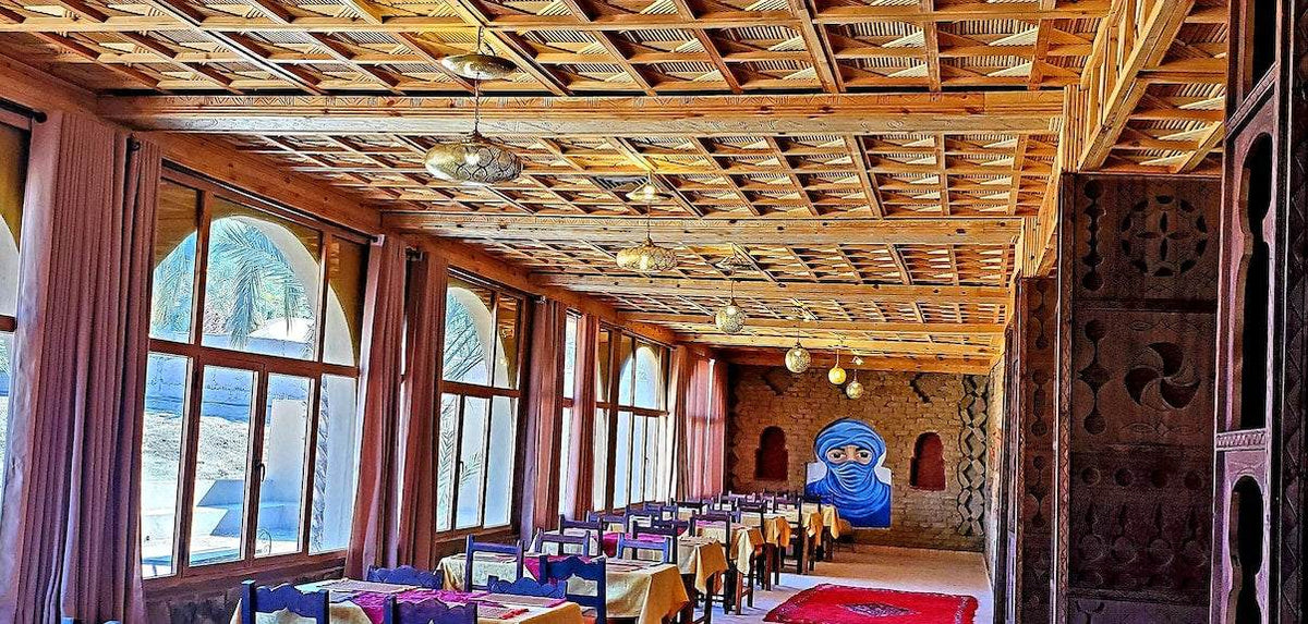 Hotel Ksar Merzouga dining room with wooden ceiling, large arched windows, and tables covered in yellow and purple tablecloths