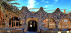 Hotel Ksar Merzouga exterior with dark stone walls, large arched windows, and wooden framed glass under a blue sky
