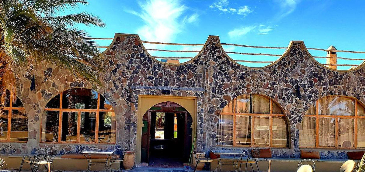 Hotel Ksar Merzouga exterior with dark stone walls, large arched windows, and wooden framed glass under a blue sky