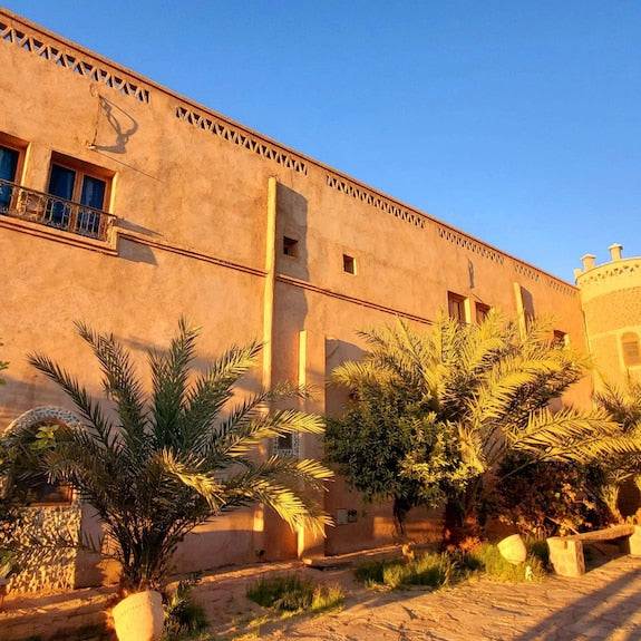 Hotel Ksar Merzouga with warm sandy-colored walls, traditional Moroccan architectural details, and lush palm trees in front under a clear blue sky