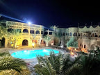 Hotel Ksar Merzouga featuring traditional beige stone architecture with arches and balconies, illuminated at night around a glowing outdoor pool surrounded by palm trees.