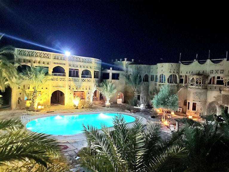 Hotel Ksar Merzouga featuring traditional beige stone architecture with arches and balconies, illuminated at night around a glowing outdoor pool surrounded by palm trees.