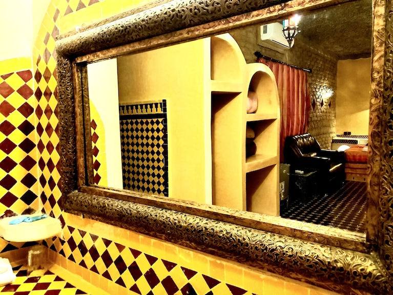 Decorative ornate gold framed mirror reflecting a cozy room with brick walls, red curtains, and dark leather chairs at Hotel Ksar Merzouga