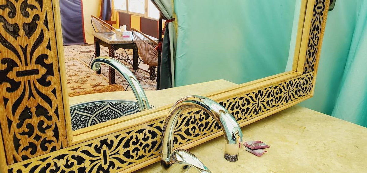 Hotel Ksar Merzouga featuring an ornate wooden framed mirror with intricate carvings above a beige marble countertop and chrome faucet