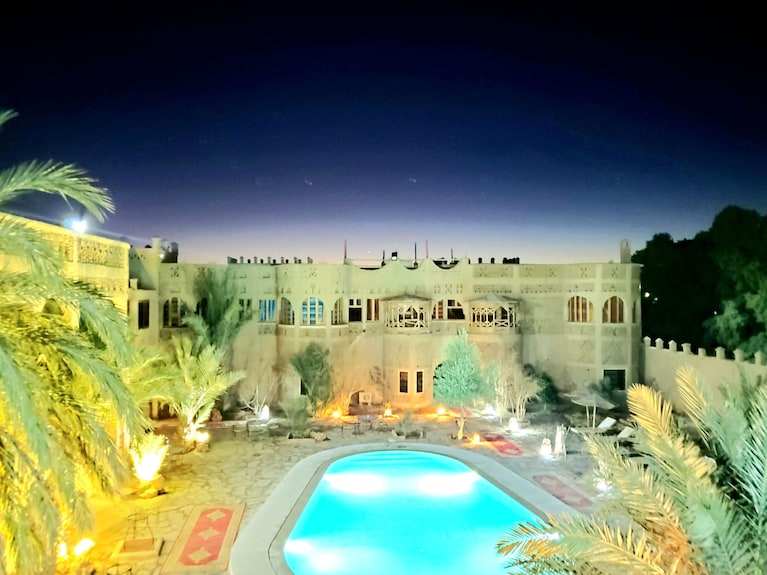 Outdoor pool glowing blue at night surrounded by desert-style architecture and palm trees at Hotel Ksar Merzouga