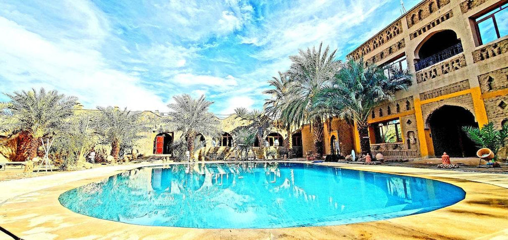 Hotel Ksar Merzouga outdoor swimming pool with blue water surrounded by palm trees and tan stone building under a blue sky