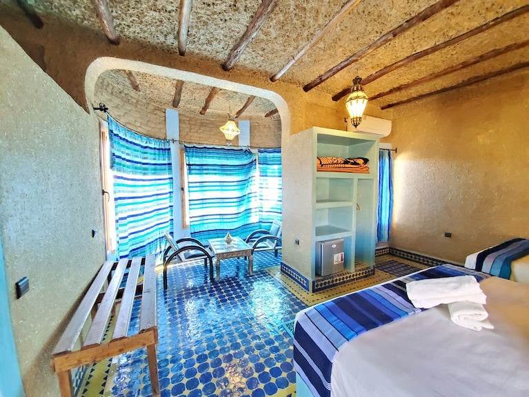 Hotel Ksar Merzouga room with textured beige walls, patterned blue tile floor, striped blue curtains, and twin beds with striped blue bedding