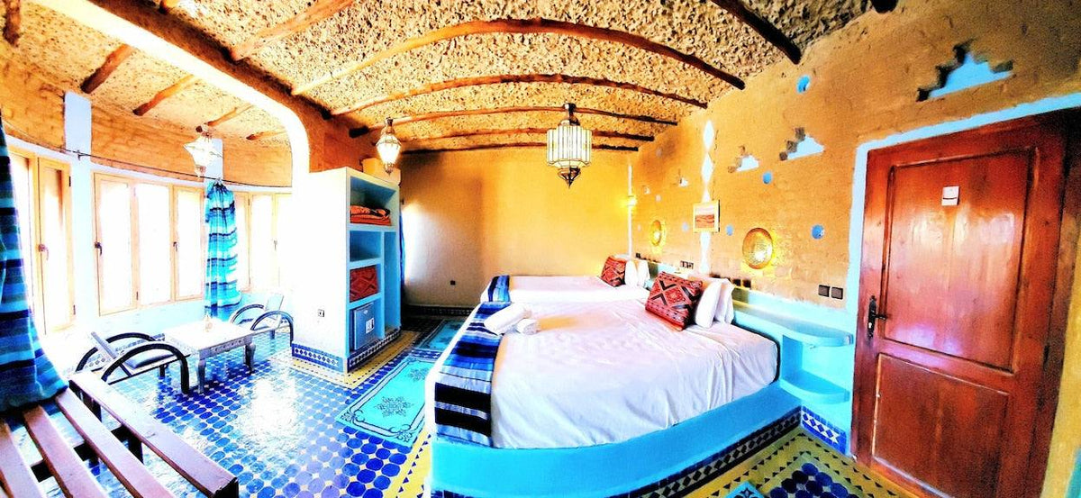 Hotel Ksar Merzouga room with twin beds, bright turquoise and yellow tiled floor, patterned cushions, and rustic yellow walls