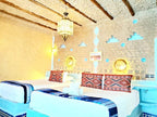 Hotel Ksar Merzouga twin beds with white linens, blue patterned throws, and decorative cushions against a rustic beige wall