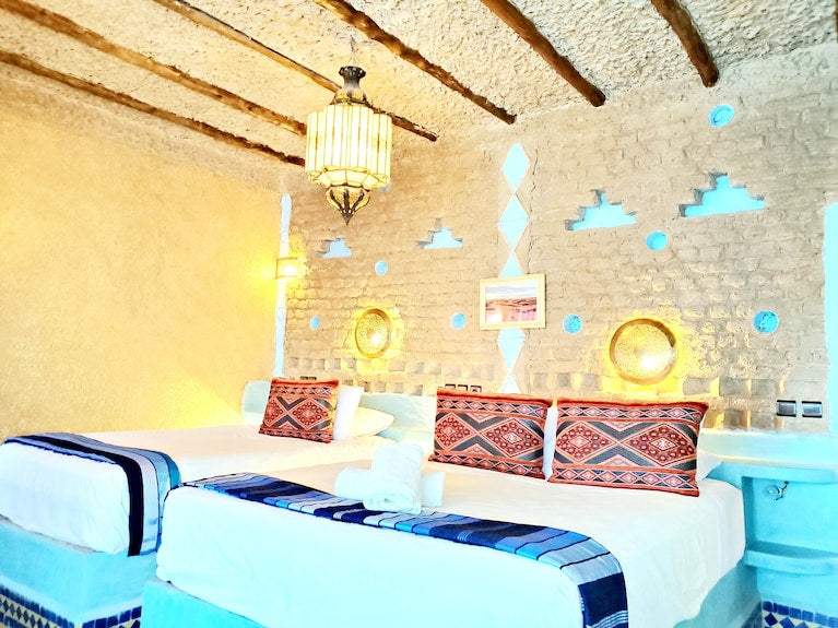 Hotel Ksar Merzouga twin beds with white linens, blue patterned throws, and decorative cushions against a rustic beige wall