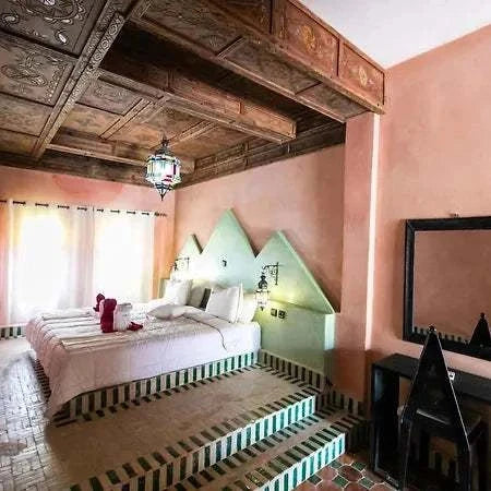 Hotel Nomad Palace bedroom with Moroccan decor featuring a white bed, geometric green patterned platform, intricate wooden ceiling, and soft peach walls