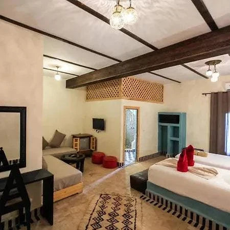Hotel Nomad Palace cozy guest room with rustic Moroccan decor, white and beige walls, wooden beams, twin beds with red towels, lounge area, and patterned rug