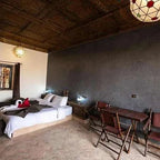 Hotel Nomad Palace room featuring a double bed with white linens, dark wooden ceiling, gray textured wall, and wooden chairs with a small table