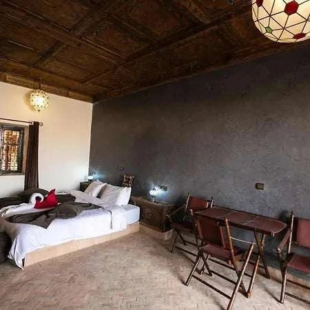 Hotel Nomad Palace room featuring a double bed with white linens, dark wooden ceiling, gray textured wall, and wooden chairs with a small table