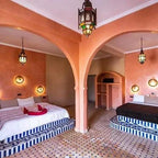 Hotel Nomad Palace room featuring two beds with striped blue and white bases, pink stucco walls, arched divider, and Moroccan lanterns