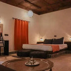 Hotel Nomad Palace bedroom with rustic wooden ceiling, warm lighting, white walls, orange curtain, and low bed with dark pillows