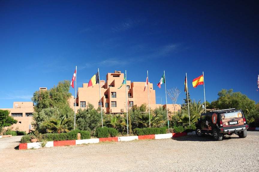 Hotel Xaluca Dades clay-colored multi-story building with flagpoles displaying various national flags and a parked black off-road vehicle
