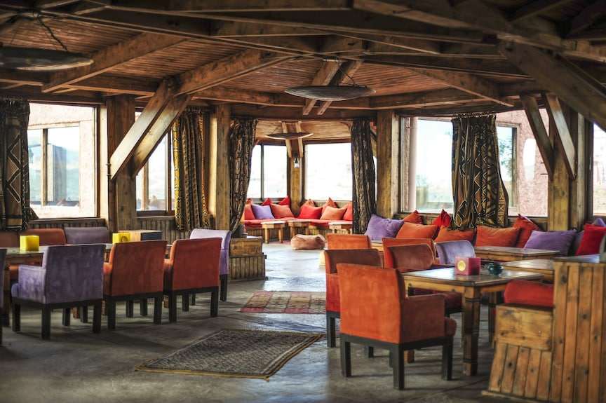 Hotel Xaluca Dades cozy lounge area with wooden beams, vibrant orange and purple chairs, patterned curtains, and rustic decor