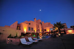 Hotel Xaluca Dades at dusk with warm desert-tone walls, illuminated outdoor lighting, swimming pool, and lounge chairs under a clear blue sky
