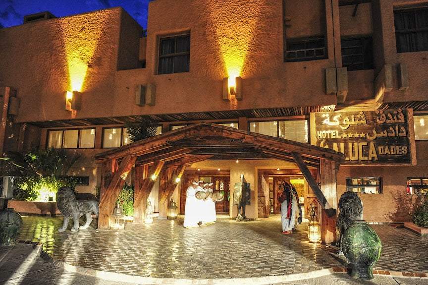 Hotel Xaluca Dades entrance at night with warm yellow lighting, traditional wooden awning, and people in white attire