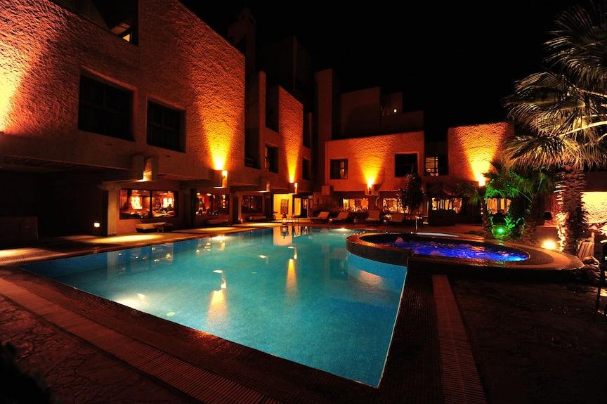 Hotel Xaluca Dades nighttime view with illuminated beige stone buildings, palm trees, and a glowing blue swimming pool
