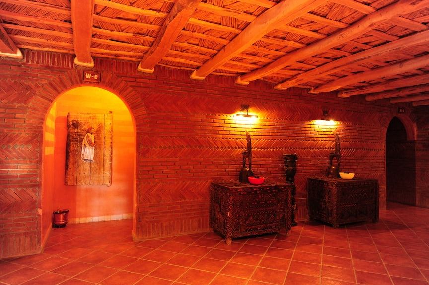 Hotel Xaluca Dades warm interior with terracotta brick walls, wooden ceiling beams, ornate dark wood chests and soft lighting