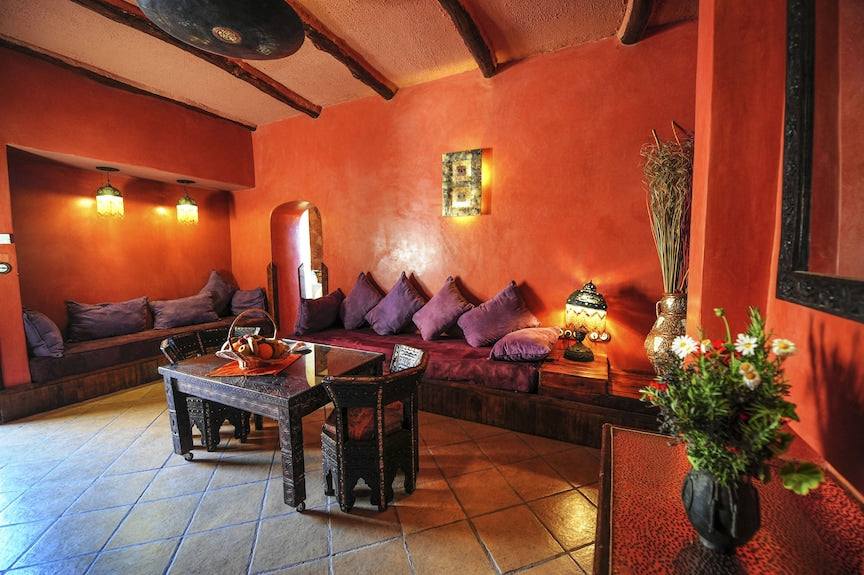 Hotel Xaluca Dades Moroccan-style lounge with warm terracotta walls, dark wooden furniture, purple cushions, and decorative lighting