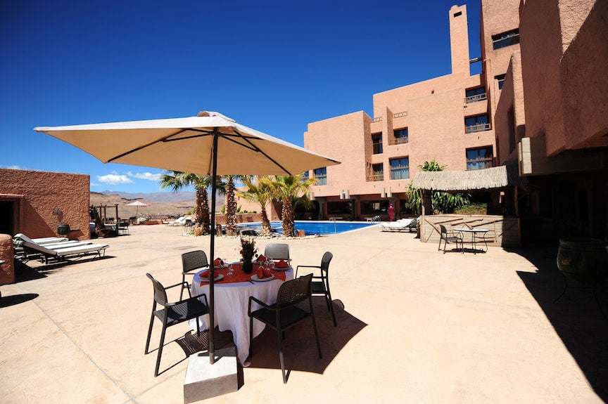 Hotel Xaluca Dades outdoor seating area with black chairs, white round table under a large beige umbrella, pool and terracotta buildings